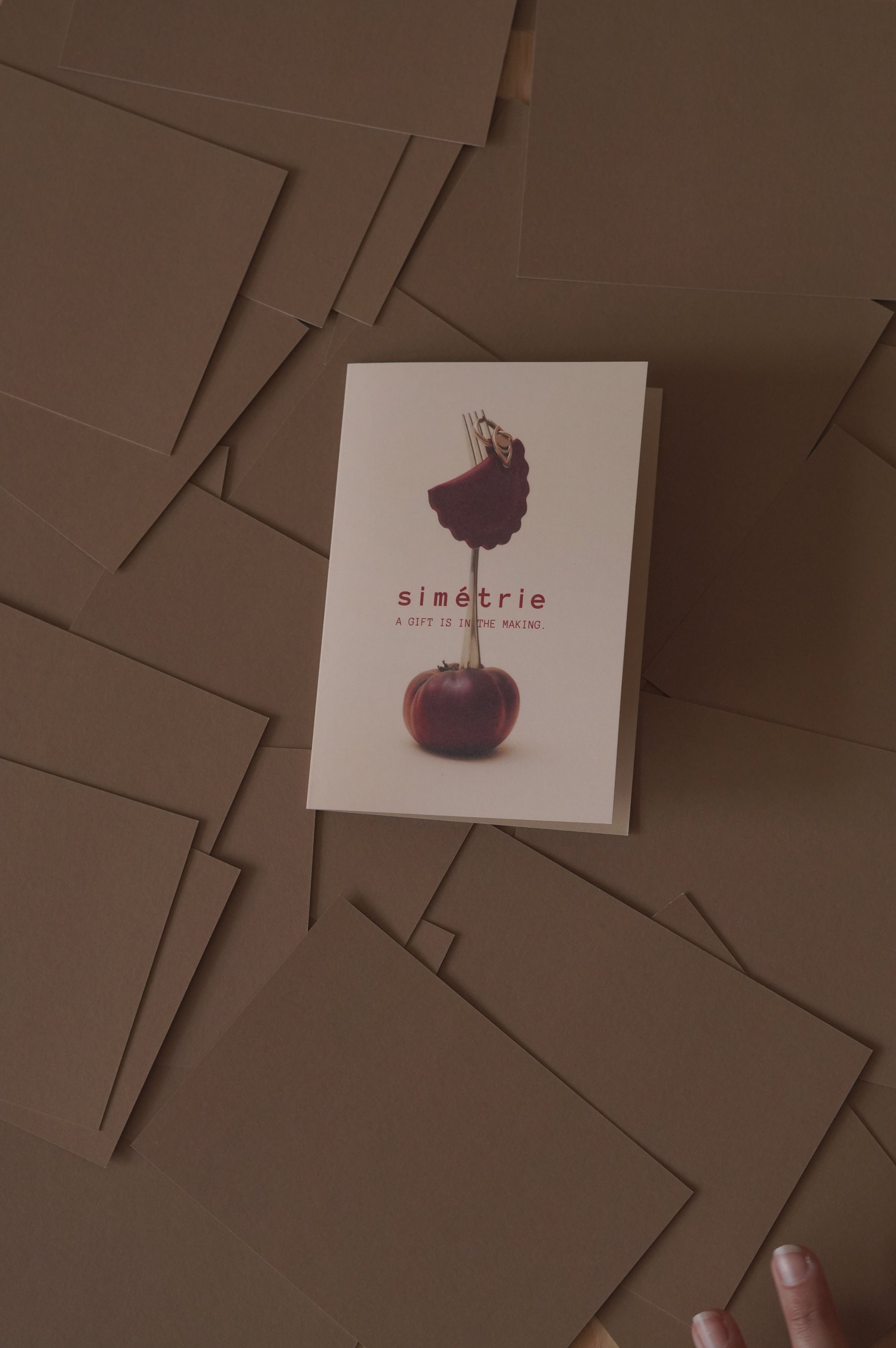 A white greeting card featuring a stylised photo of a dark tomato and the text 'simétrie', placed on top of a scattered pile of brown envelopes.