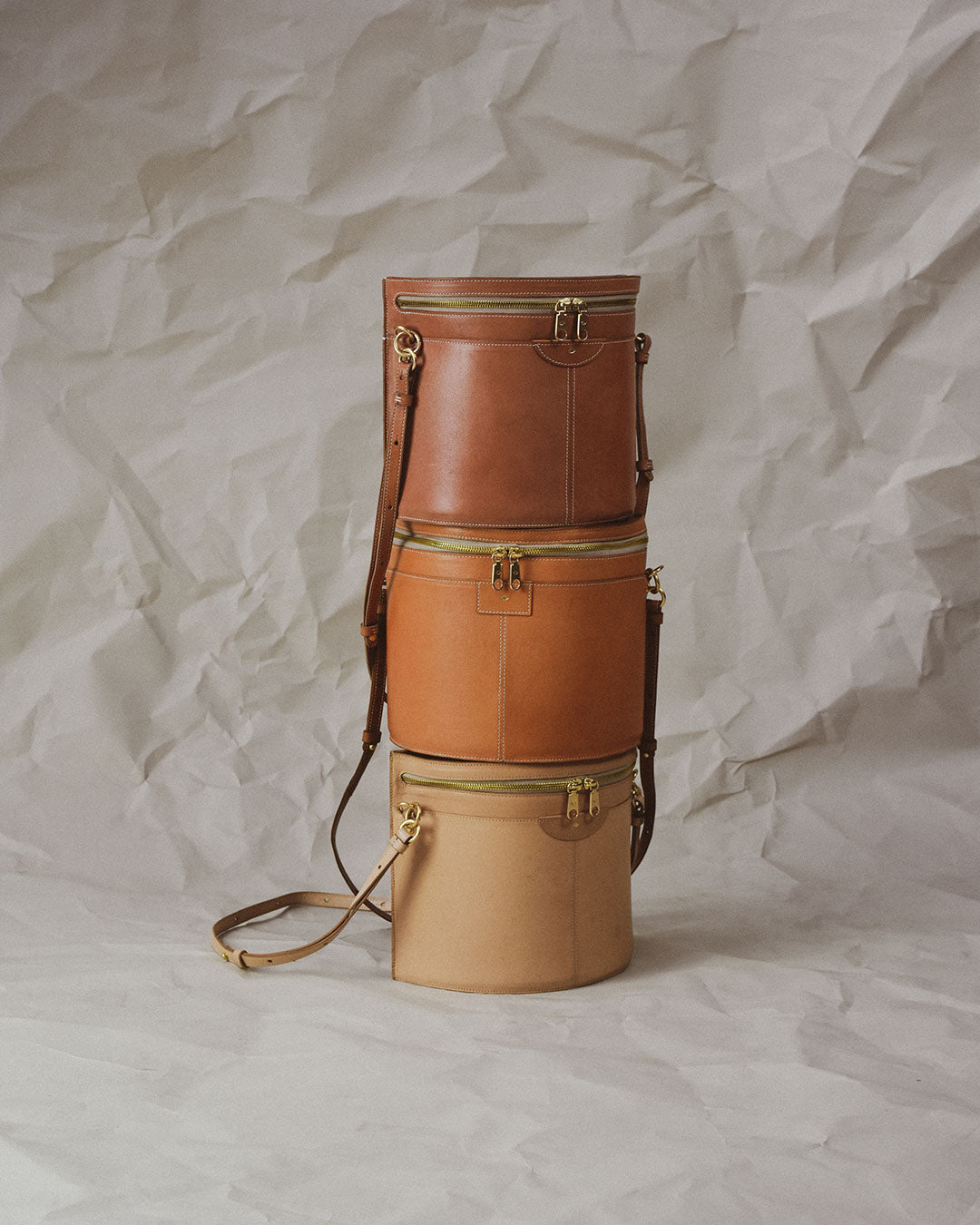 Three pastel coloured leather bucket bags are stacked on top of eachother. They are centered within the image, with moody lighting, and a crumpled paper background.