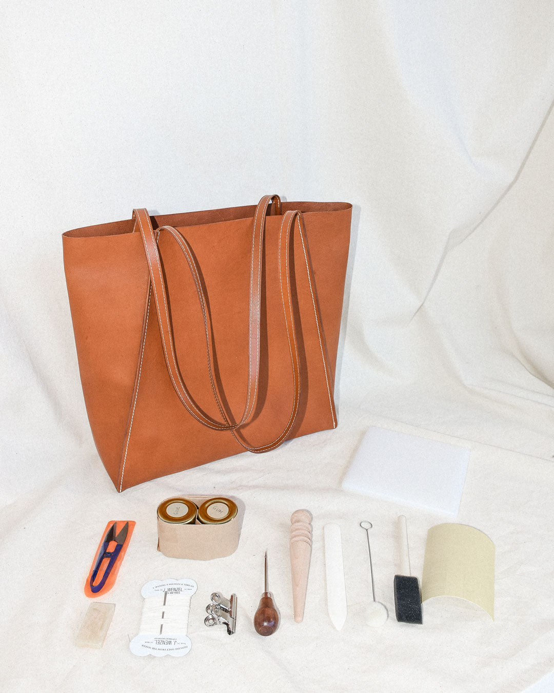 A tan leather tote bag is the focal point of the image. Various leather tools are in the foreground, such as an awl, snips, and a burnishing rod.