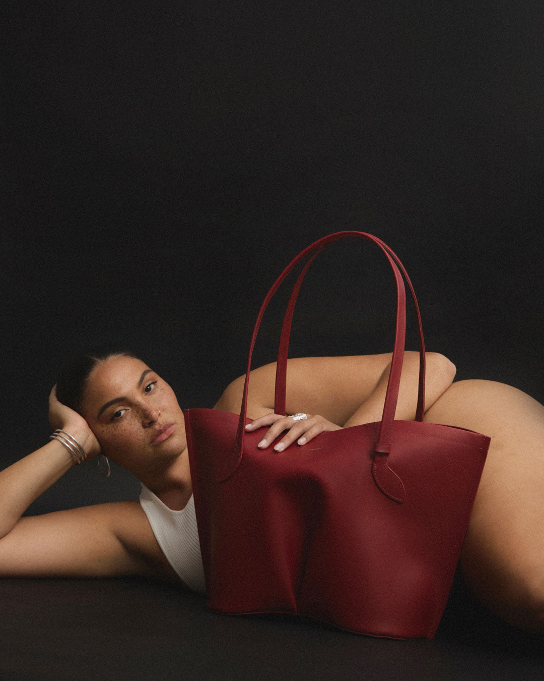 A woman, Caitlin Murphy, laying on a dark surface beside a large structured leather tote bag in deep burgundy red. The bag has two long handles and a minimalist silhouette, photographed against a dark studio background.