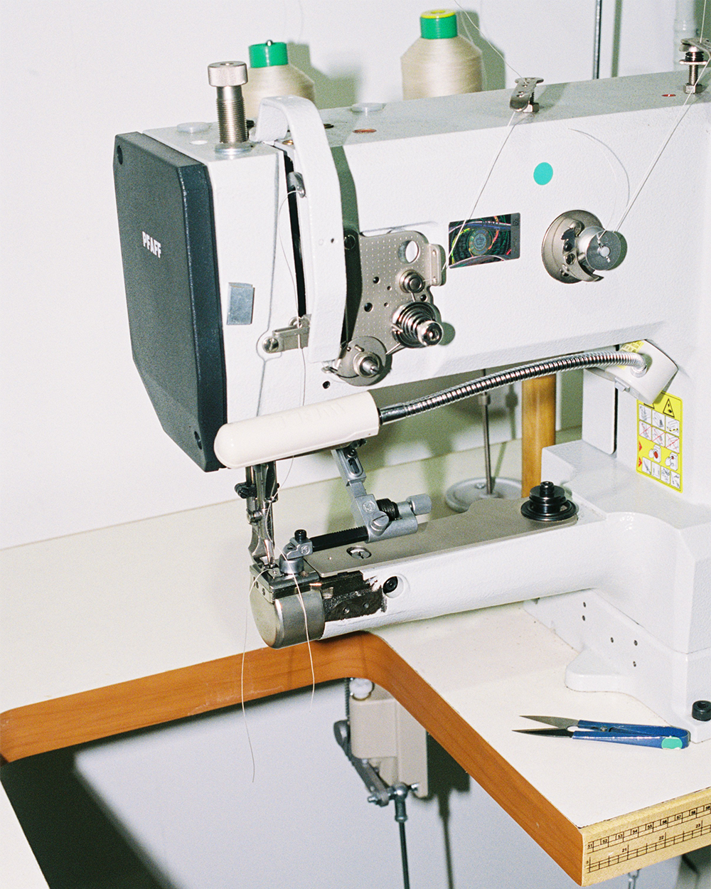 Inside the simétrie atelier: Close-up of a professional industrial sewing machine in a workspace. The machine is branded "Pfaff" and is set up with spools of beige and green thread on top. Various mechanical components, including levers, spools, and tension knobs, are visible. The corner of a wooden workbench is seen in the foreground, along with a small pair of blue-handled scissors resting on the surface.