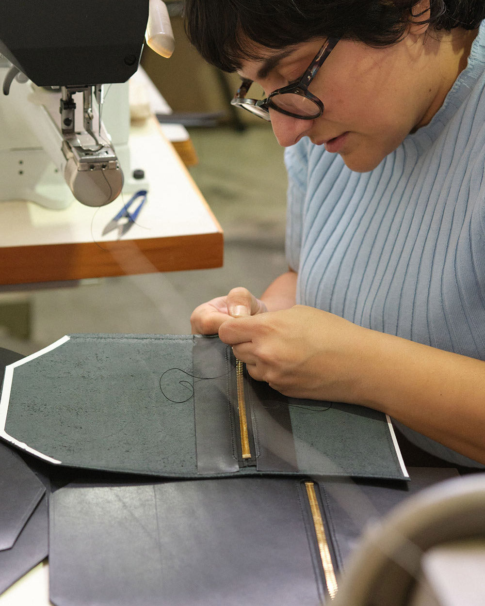 A close up of Simone at the sewing machine. She has her head down and is tying off the threads on the leather bag panel she was sewing.