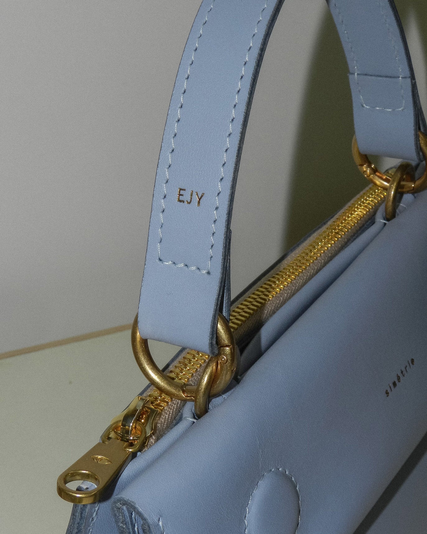 Close-up view of a light blue leather handbag strap. The strap is attached to the bag with gold-coloured hardware. On the strap, there are visible stitches along the edges and the initials "EJY" are embossed in gold lettering. Part of the bag's zipper, also in gold colour, is visible. The background is a neutral off-white colour.